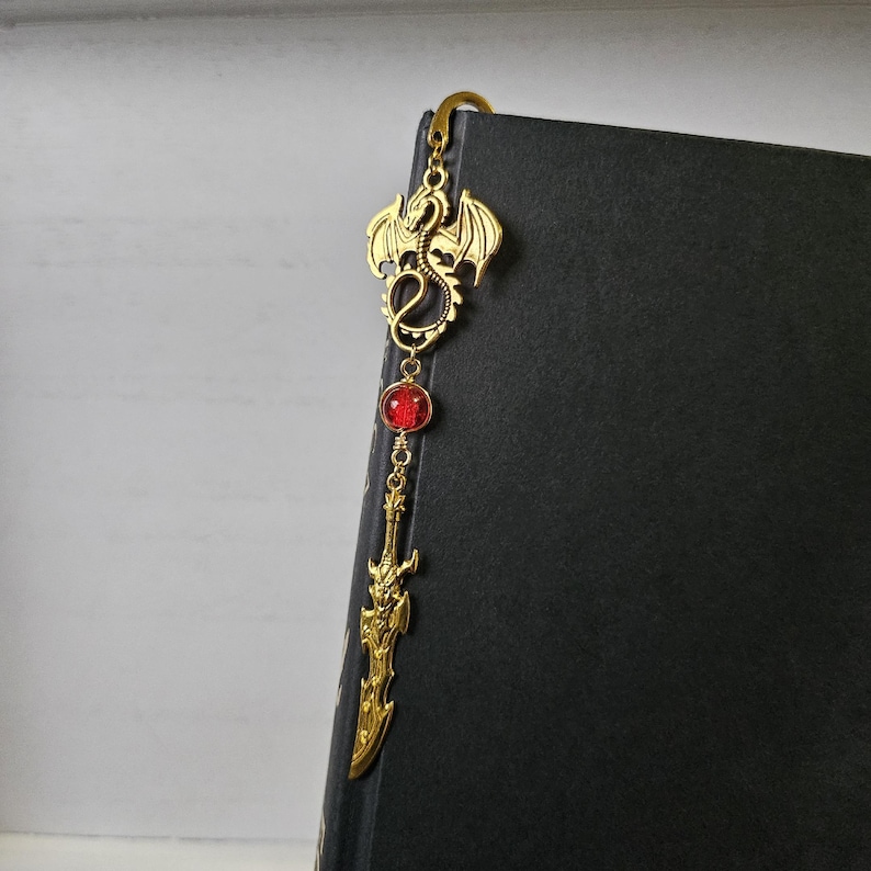 May include: Gold bookmark featuring a dragon design with a red gemstone and a sword pendant.