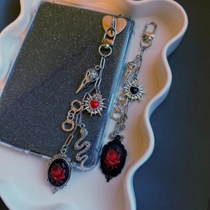May include: Two gothic-style phone charms with silver chains and decorative elements. One charm features a raven skull, a red heart, and a snake. The other has a sunburst, a black heart, and a cameo with a red rose.