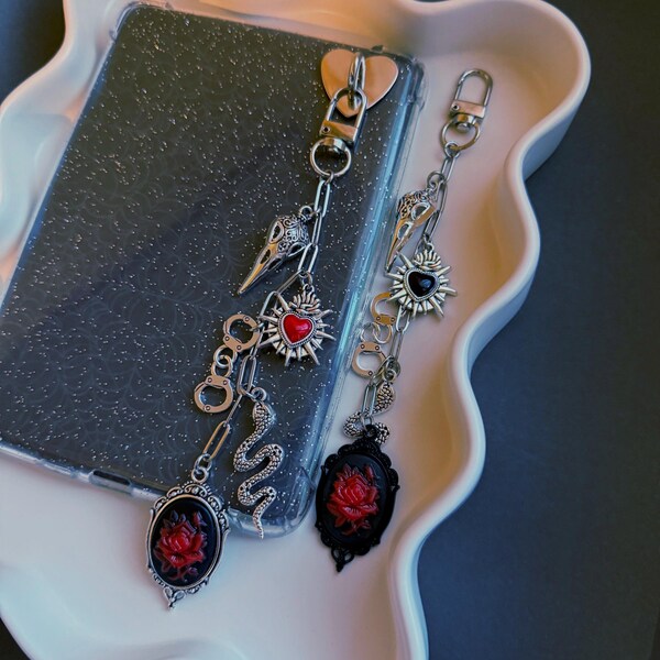 Puede incluir: Dos colgantes góticos para teléfono con cadenas plateadas y elementos decorativos. Uno presenta una calavera de cuervo, un corazón rojo y una serpiente. El otro tiene un sol, un corazón negro y un camafeo con una rosa roja.