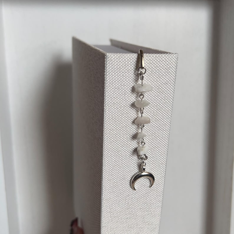May include: A silver crescent moon charm hangs from a silver chain with white stones. The chain is attached to a book.