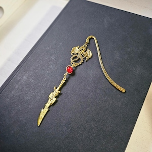 May include: Gold bookmark featuring a dragon design with a red gemstone. The bookmark is shaped like a sword and has a decorative, ornate design.