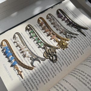 May include: A set of ten gold and silver bookmark charms with various gemstone accents. The charms feature a crescent moon, a sun, a dragon, and a star. The gemstones are in shades of blue, green, pink, and purple.
