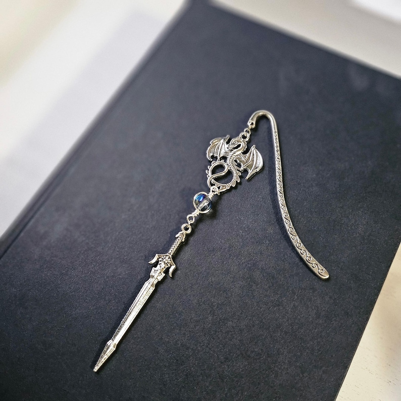 May include: Silver bookmark featuring a dragon design with a sword and a blue iridescent bead. The bookmark is on a dark gray surface.