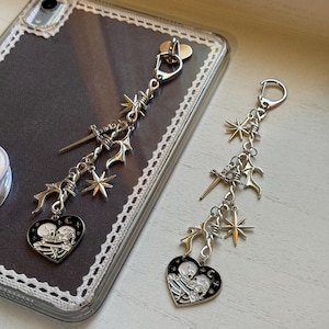 May include: Silver-toned phone charms with a gothic aesthetic. Each charm features a heart-shaped pendant with a skeleton couple, a dagger, starbursts, and other decorative elements. The charms are attached to a silver clasp.