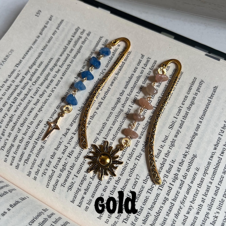 May include: Two gold bookmark designs with decorative edges. One bookmark features a sun with a gold finish and the other has a crystal charm with blue and pink stones. Both bookmarks have a gold chain.