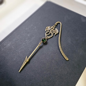 May include: A gold dragon bookmark with a green gemstone. The bookmark has a long, thin, pointed end and a decorative, curled tail.