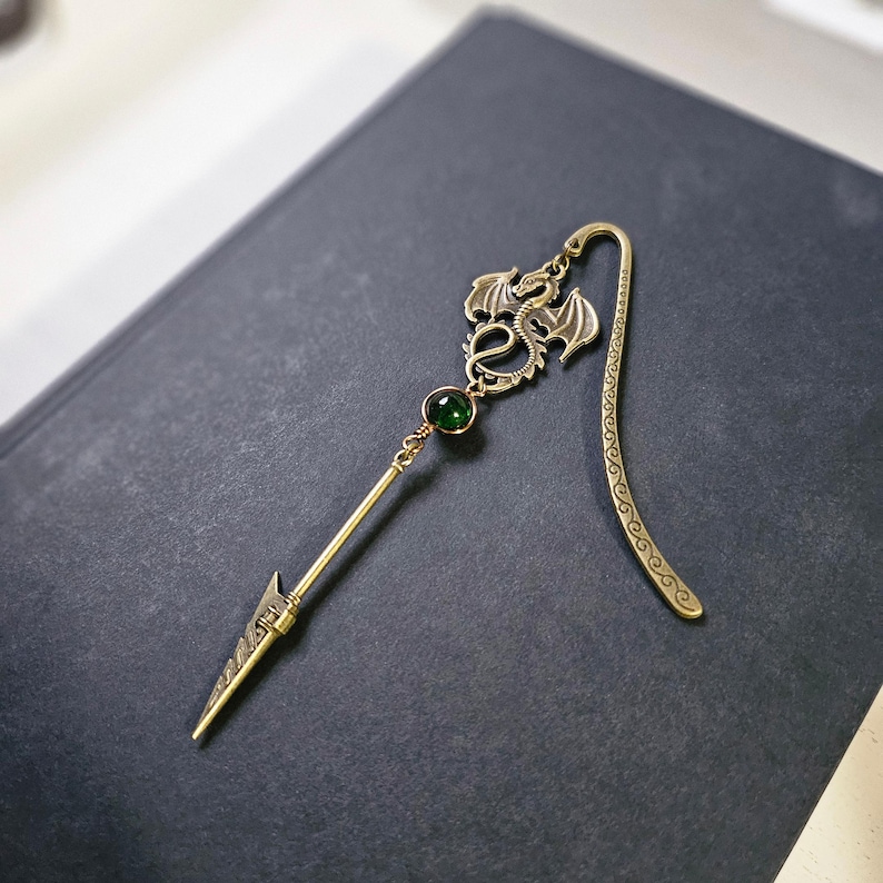 May include: A gold dragon bookmark with a green gemstone. The bookmark has a long, thin, pointed end and a decorative, curled tail.