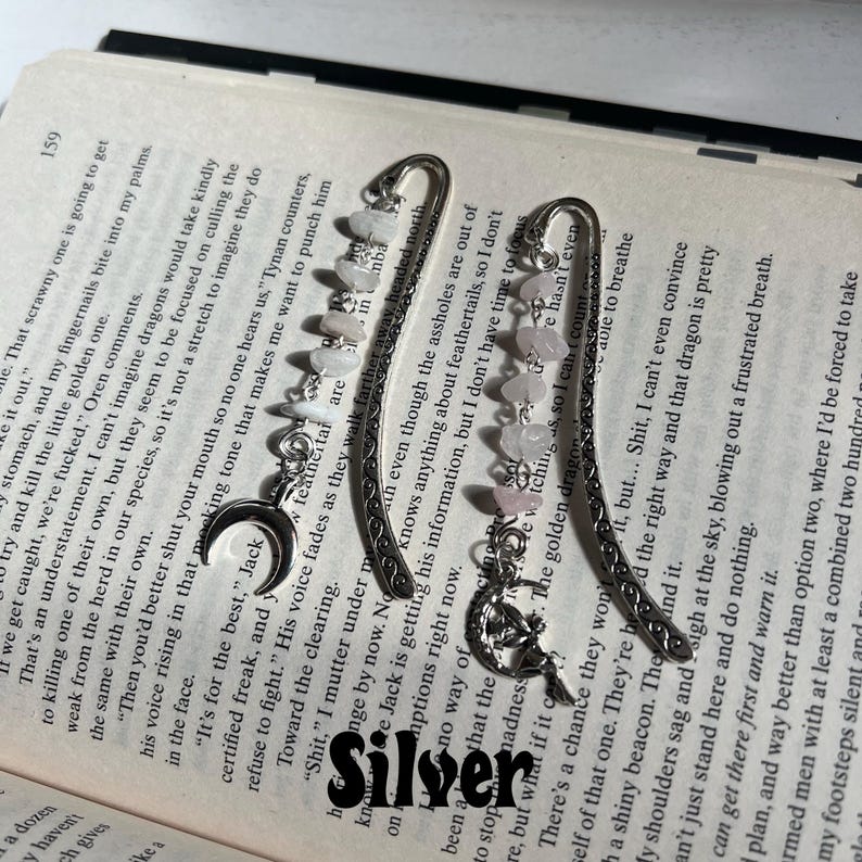 May include: Two silver bookmark with a crescent moon charm and pink and white stones. The bookmarks are decorated with intricate designs.