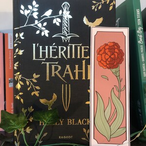 Peut inclure: Un livre noir avec des lettres dorées qui lisent "L'Héritage Trahi" par Holly Black. Le livre est sur une étagère en bois avec une plante verte devant. Un marque-page rose avec une illustration de fleur rouge dépasse du livre.