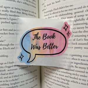 Puede incluir: Un marcapáginas en forma de burbuja de diálogo, con un efecto acuarela rosa y azul, en el que se lee el texto "The Book Was Better" en letras negras. El marcapáginas está colocado en una página de un libro con texto sobre un personaje llamado Huck.