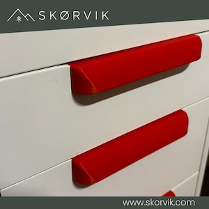 May include: A white storage unit with multiple drawers featuring bright red, horizontal, rectangular handles. The top of the unit displays the text SKØRVIK with a mountain icon, and the bottom edge includes the website text www.skorvik.com.