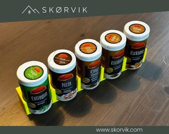 May include: A bright yellow plastic spice rack holding five cylindrical Ostmann spice containers. The labels on the containers read Estragon, Pizza, Kreuzkümmel, Koriander, and Kurkuma. The rack is designed to hold each container upright.