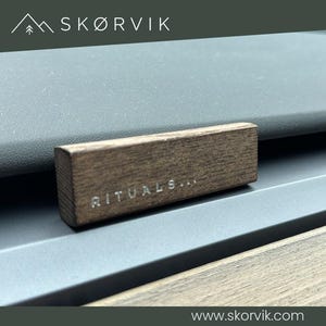 Könnte beinhalten: Ein rechteckiger, dunkelbrauner Holz-Clip für die Autolüftung mit sichtbarer Holzmaserung. Das Wort RITUALS... ist in weißen Buchstaben auf die Vorderseite des Holzblocks eingraviert, der an einer grauen horizontalen Lüftungslamelle befestigt ist.