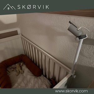 May include: A white baby cot featuring a white adjustable camera mount attached to the side rail. The cot contains a rust-coloured fabric bumper and a patterned blanket with a small plush toy. The SKØRVIK logo is visible at the top of the image.