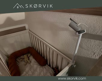 May include: A white baby cot featuring a white adjustable camera mount attached to the side rail. The cot contains a rust-coloured fabric bumper and a patterned blanket with a small plush toy. The SKØRVIK logo is visible at the top of the image.