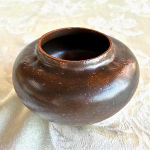 May include: A small, round, dark brown ceramic bowl with a smooth, glossy finish and a wide, open top. The vessel features a slightly textured surface with subtle variations in color, showcasing a deep, earthy tone and a rounded, squat shape.