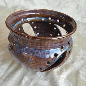 May include: A round, glazed ceramic bowl featuring a deep brown and blue mottled finish. The vessel has a wide, open top and is punctuated by several large, curved cutouts along the sides, accented by a series of small, evenly spaced circular holes.