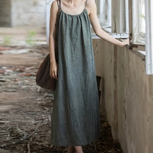 May include: A sage green maxi dress with thin spaghetti straps. The dress has a gathered bodice and a flowing skirt. The fabric appears to be a lightweight linen or cotton blend. A large brown shoulder bag is also visible.