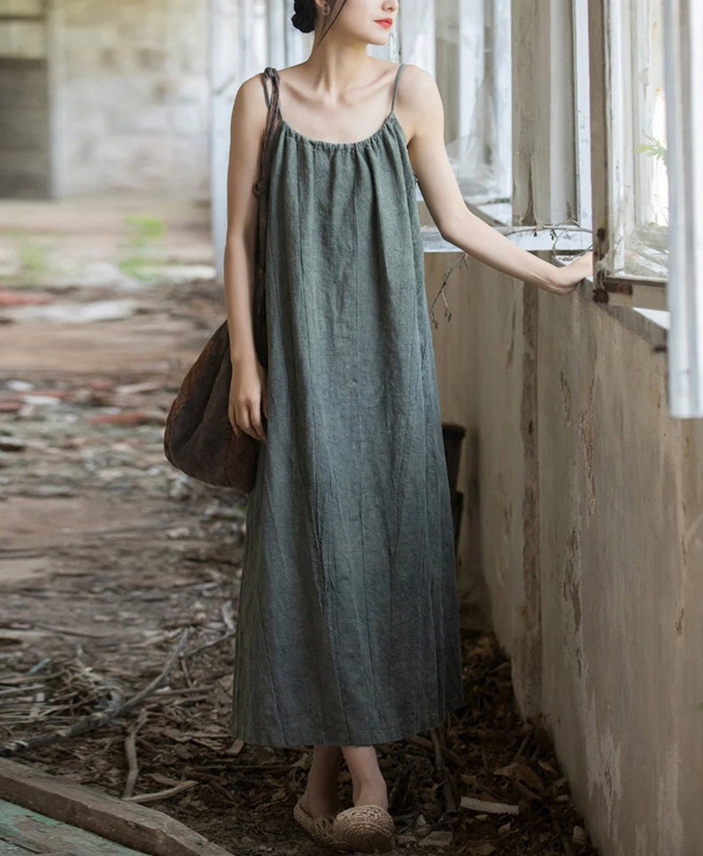 May include: A sage green maxi dress with thin spaghetti straps. The dress has a gathered bodice and a flowing skirt. The fabric appears to be a lightweight linen or cotton blend. A large brown shoulder bag is also visible.