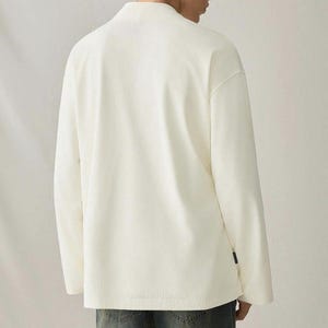 May include: Cream-colored long-sleeve shirt with a mandarin collar. The shirt has a relaxed fit and a minimalist design. The model is wearing blue jeans. The shirt has a small black tag on the lower left side.