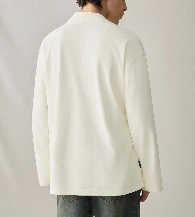May include: Cream-colored long-sleeve shirt with a mandarin collar. The shirt has a relaxed fit and a minimalist design. The model is wearing blue jeans. The shirt has a small black tag on the lower left side.