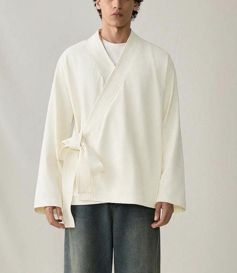 May include: Cream-colored wrap top with long sleeves and a tie closure. The top has a V-neck design and is paired with blue denim jeans. The fabric appears to be a soft, lightweight material.