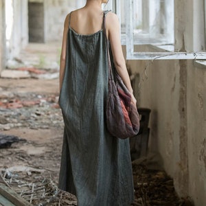 May include: A long, olive green dress with thin straps. The dress has a textured appearance and falls to the ankles. A patterned shoulder bag in shades of red, brown, and orange is worn over the shoulder. The setting appears to be an old building.