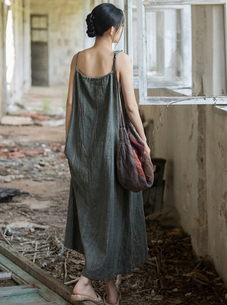 May include: A long, olive green dress with thin straps. The dress has a textured appearance and falls to the ankles. A patterned shoulder bag in shades of red, brown, and orange is worn over the shoulder. The setting appears to be an old building.