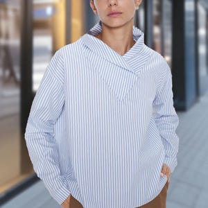 May include: A light blue and white striped long-sleeve shirt with a unique collar design. The shirt is paired with brown pants. The model is standing in front of a blurred background.