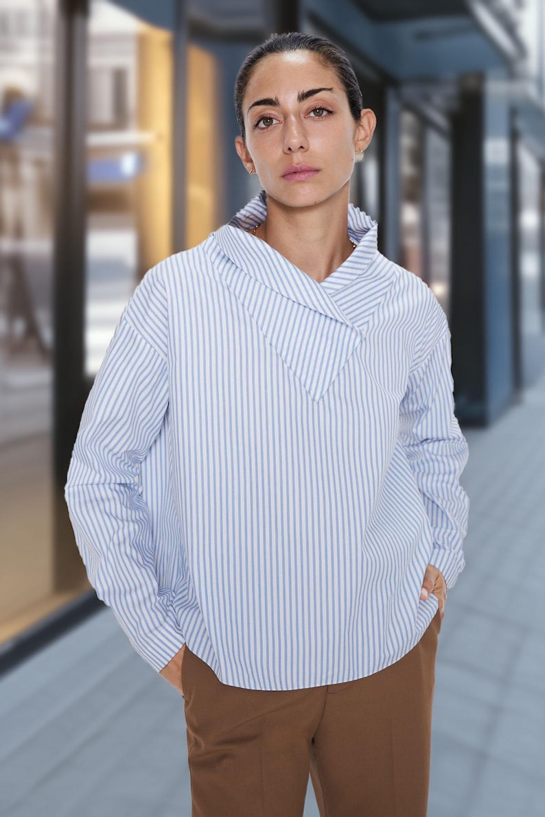 May include: A light blue and white striped long-sleeve shirt with a unique collar design. The shirt is paired with brown pants. The model is standing in front of a blurred background.