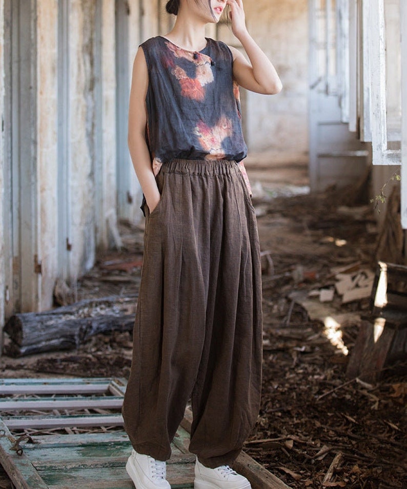 May include: A woman wearing a brown sleeveless top with a floral print and brown wide-leg pants. She is standing in a room with a wooden floor and white walls.