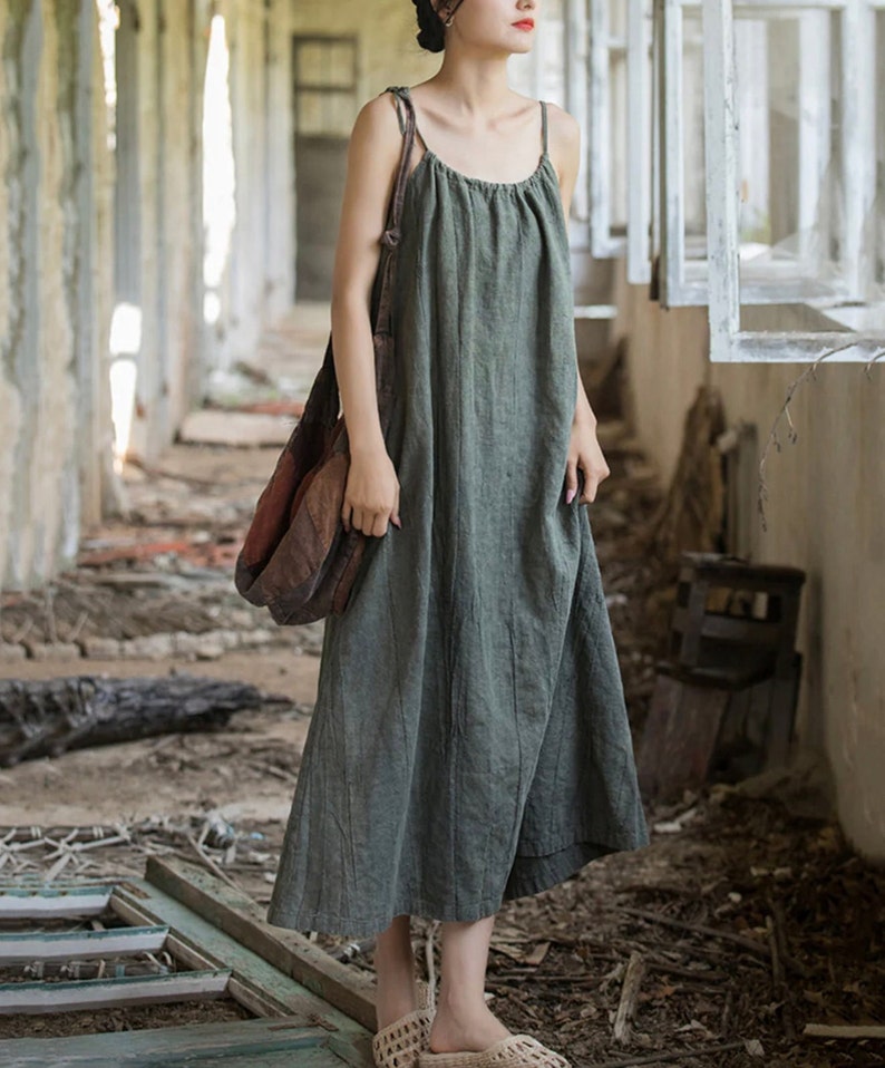 May include: A sage green, sleeveless dress with thin straps. The dress has a loose, flowing fit and reaches mid-calf. A brown shoulder bag is worn over the shoulder. The dress is made of a textured fabric.