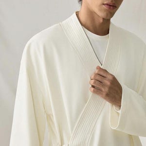 May include: Cream-colored robe with a wrap-around design. The robe features a textured collar and lapel, and a matching belt. A silver chain necklace is visible. The robe is made of a soft, flowing fabric.