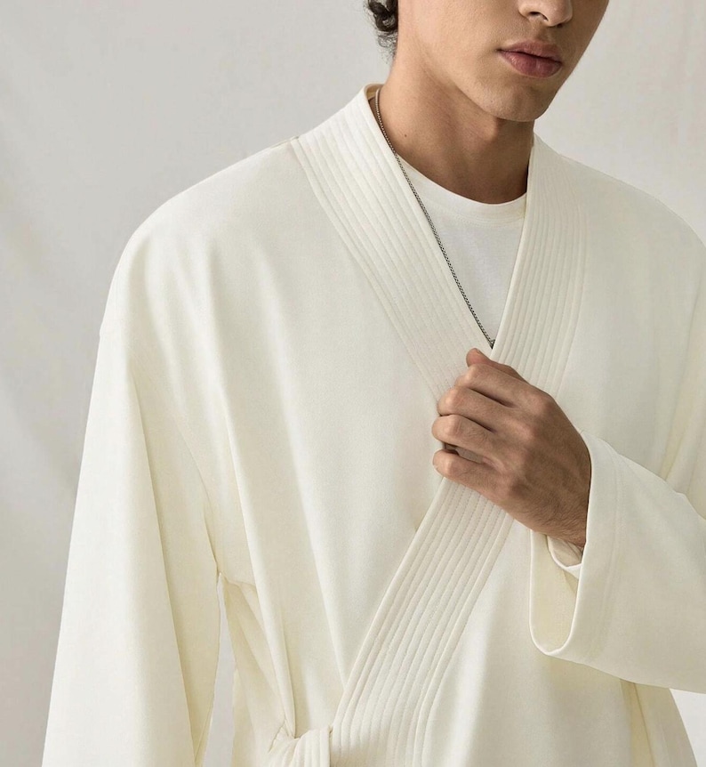 May include: Cream-colored robe with a wrap-around design. The robe features a textured collar and lapel, and a matching belt. A silver chain necklace is visible. The robe is made of a soft, flowing fabric.