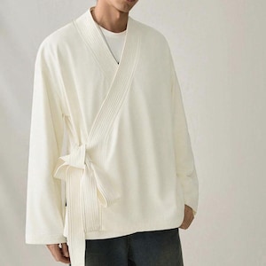 May include: Cream-colored wrap top with long sleeves and a tie closure. The top features a ribbed design on the front and a relaxed fit. The garment is made of a soft, lightweight fabric, suitable for layering or wearing on its own.