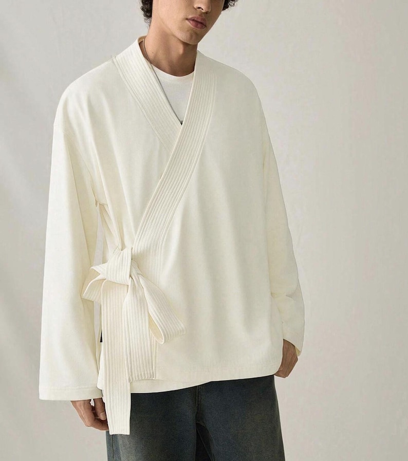 May include: Cream-colored wrap top with long sleeves and a tie closure. The top features a ribbed design on the front and a relaxed fit. The garment is made of a soft, lightweight fabric, suitable for layering or wearing on its own.