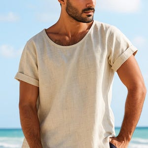 May include: Beige linen-blend t-shirt with a wide, rounded neckline and rolled-up short sleeves. The shirt has a relaxed fit and a textured appearance. The model is wearing navy blue shorts. The background is a beach scene.