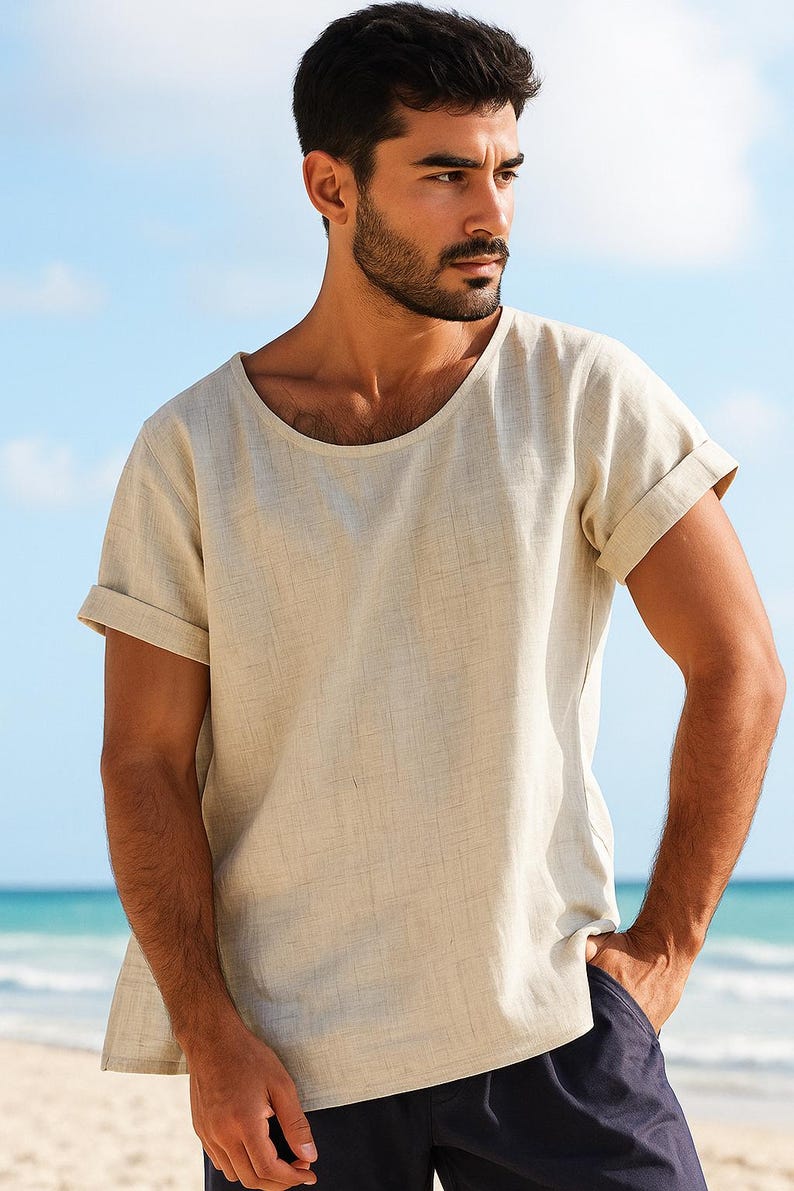 May include: Beige linen-blend t-shirt with a wide, rounded neckline and rolled-up short sleeves. The shirt has a relaxed fit and a textured appearance. The model is wearing navy blue shorts. The background is a beach scene.