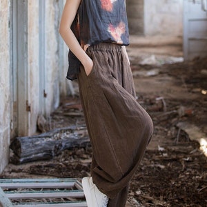 May include: A woman wearing a brown linen pants with a loose fit and a black sleeveless top with a tie-dye pattern. She is wearing white sneakers and standing on a wooden surface.