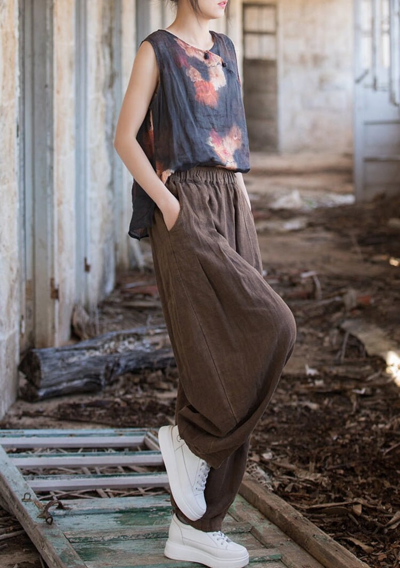 May include: A woman wearing a brown linen pants with a loose fit and a black sleeveless top with a tie-dye pattern. She is wearing white sneakers and standing on a wooden surface.