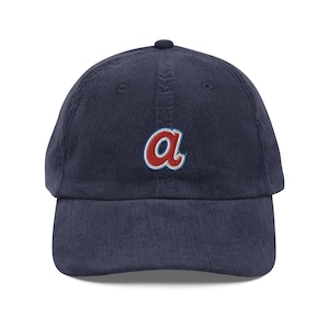 May include: A navy blue corduroy baseball cap with a red and white embroidered "A" on the front.