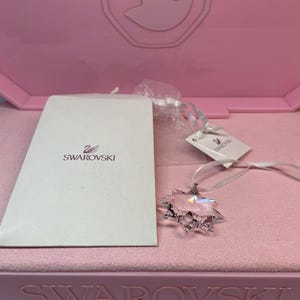 Puede incluir: Un colgante de estrella de cristal transparente con cierre plateado y cinta blanca, exhibido sobre una superficie de terciopelo rosa. El colgante está junto a una tarjeta blanca con el logotipo de Swarovski y una caja rosa.