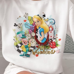 May include: White sweatshirt with a vibrant illustration of Alice in Wonderland characters, including Alice, the Mad Hatter, and the Cheshire Cat. The text "Alice in Wonderland" is also displayed. The sweatshirt is worn by a person.