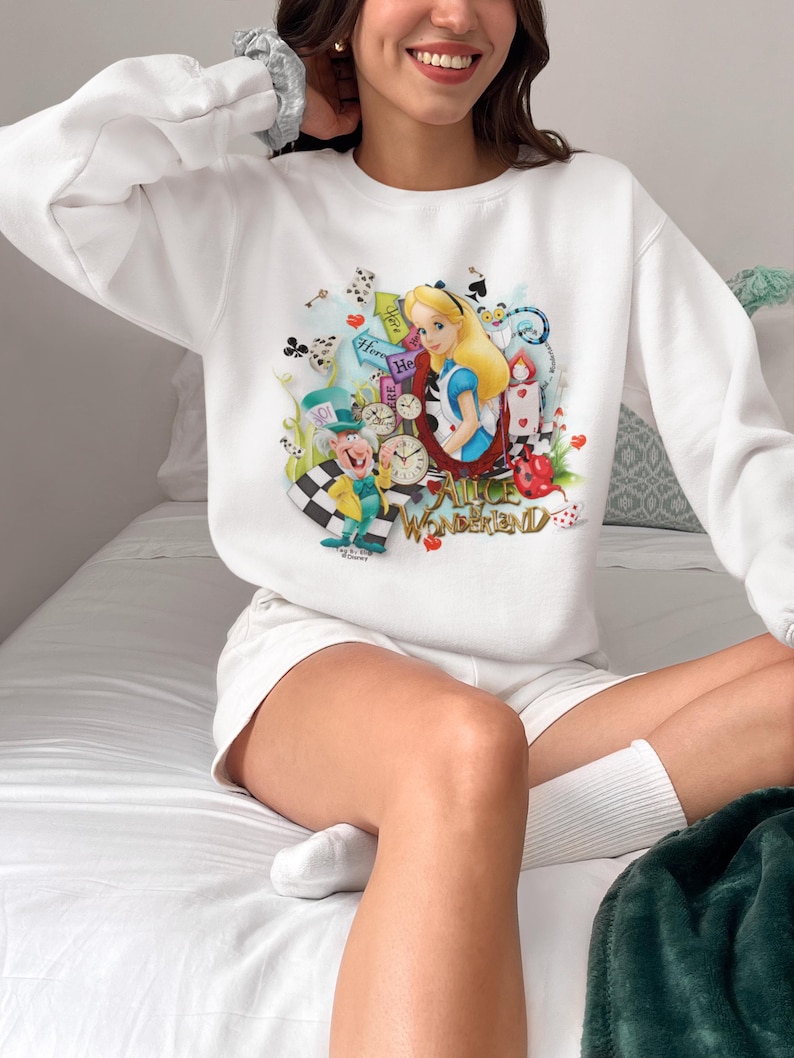 May include: White sweatshirt with a vibrant illustration of Alice in Wonderland characters, including Alice, the Mad Hatter, and the Cheshire Cat. The text "Alice in Wonderland" is also displayed. The sweatshirt is worn by a person.