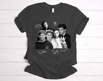 May include: A dark gray t-shirt with a black and white photo of five men. The men are all wearing different styles of clothing. The photo is surrounded by white signatures. The signatures are "Harry", "Liam", "Zayn", "Niall", and "Louis".