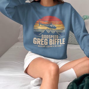 May include: A blue crew neck sweatshirt with a graphic of a helicopter, mountains, and a sunset. The text reads "GODSPEED GREG BIFFLE - NASCAR LEGEND & NC HERO." The sweatshirt is a casual style.