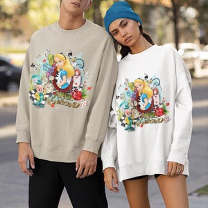 May include: Two sweatshirts, one beige and one white, with a vibrant Alice in Wonderland design. The illustration includes Alice, clocks, playing cards, and the words "Alice in Wonderland". The beige sweatshirt is worn with black trousers, the white one with black shorts.