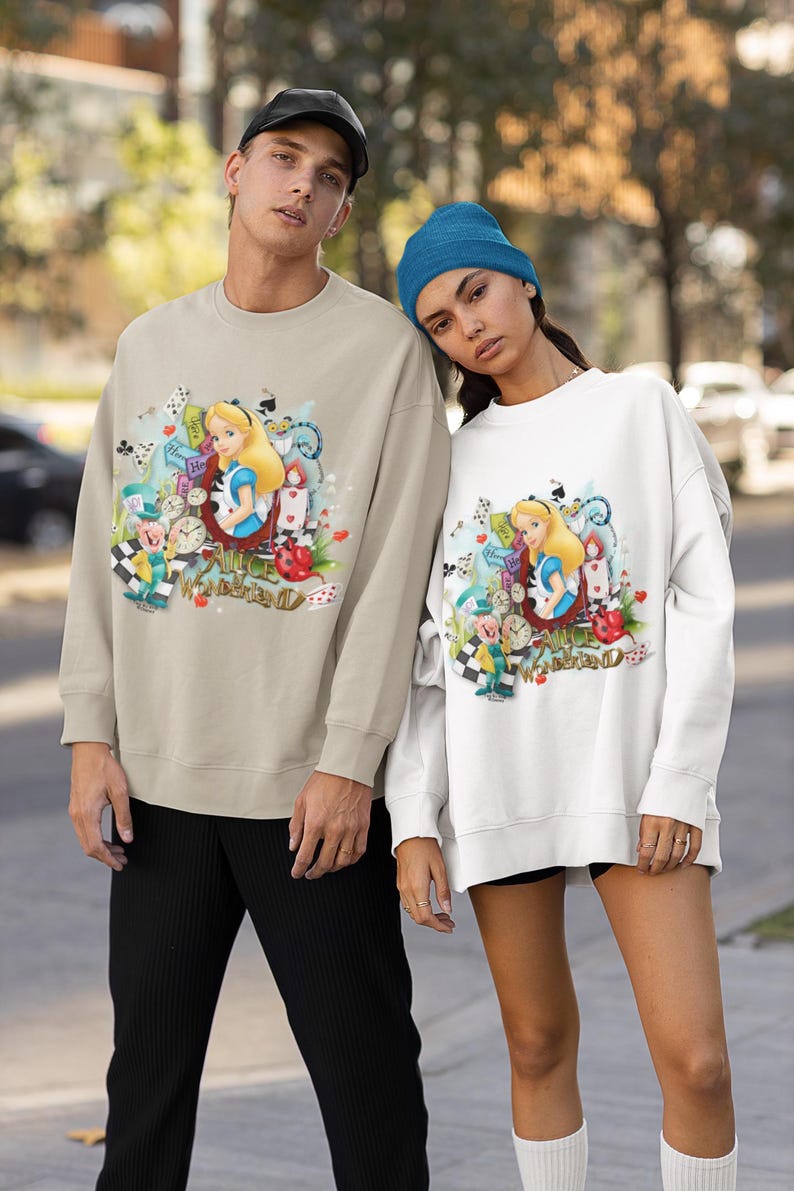 May include: Two sweatshirts, one beige and one white, with a vibrant Alice in Wonderland design. The illustration includes Alice, clocks, playing cards, and the words "Alice in Wonderland". The beige sweatshirt is worn with black trousers, the white one with black shorts.