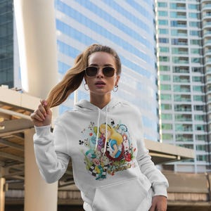 May include: White hoodie with a colourful Alice in Wonderland illustration. The design features Alice, the Mad Hatter, and other characters from the story. The sweatshirt has a front pocket and a drawstring hood.