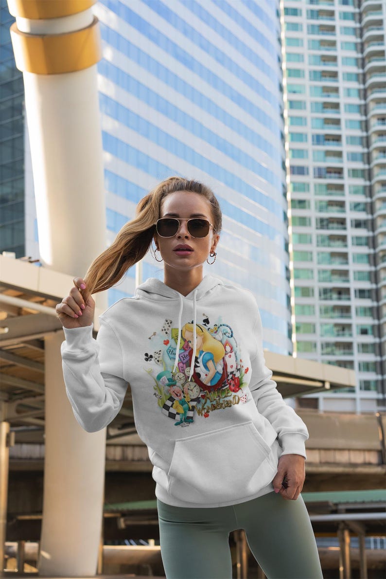 May include: White hoodie with a colourful Alice in Wonderland illustration. The design features Alice, the Mad Hatter, and other characters from the story. The sweatshirt has a front pocket and a drawstring hood.